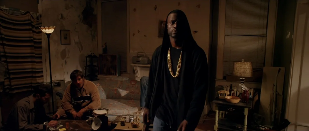 Rage (2014) movie still: medium — A cluttered apartment living room features peeling wallpaper and old wooden furniture pieces. A man in a black…