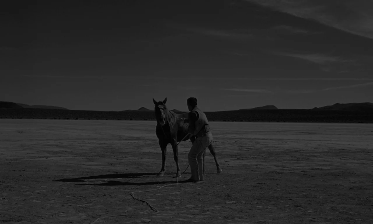 The Misfits (1961) movie still: wide — A vast, flat desert landscape stretches out under a dark, minimal sky. A man stands holding a rope connected…