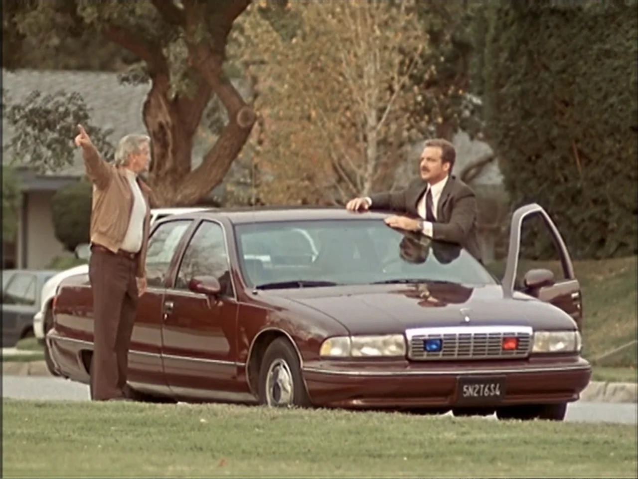 Just 4 Kicks (2003) movie still: fullbody — A dark red sedan sits on a residential street lined with green grass and trees. Two men stand beside the…
