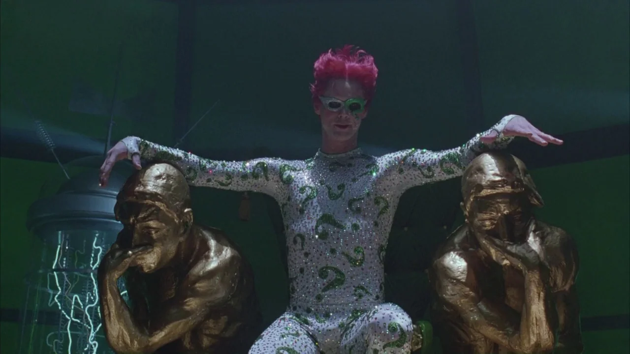 Batman Forever (1995) movie still: medium — A dark, green-lit room featuring metallic gold statues and complex electrical machinery. A slender man…