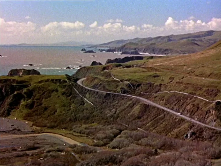 The Birds movie still: establishing — A winding road cuts through grassy hills overlooking a vast ocean bay. Small white clouds drift above distant,…