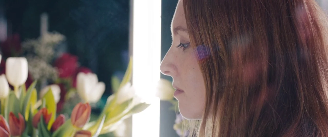 Kiss of the Damned (2012) movie still: closeup — A side profile of a young woman with auburn hair stands near a bouquet. Bright white light from a…