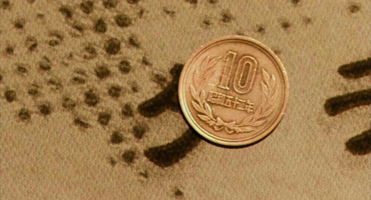 Stigmatized Properties: Possession (2025) movie still: closeup — A Japanese ten-yen bronze coin lies flat on a light brown textured surface. Dark…