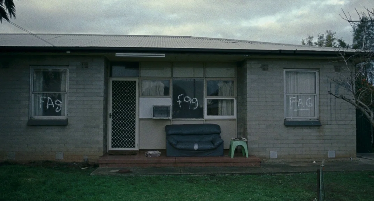 Snowtown (2011) movie still: establishing — A single-story brick house with a dark sofa placed on the front porch. The windows are tagged with…
