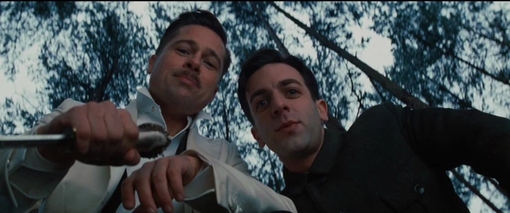 Inglourious Basterds (2009) movie still: medium — Two men stand over the camera in a densely wooded area with tall trees. They stare directly…