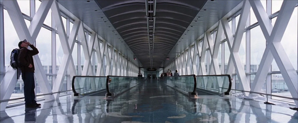 The Departed (2006) movie still: wide — A long, enclosed modern glass walkway features an elevated moving walkway in the center. A man stands at the…