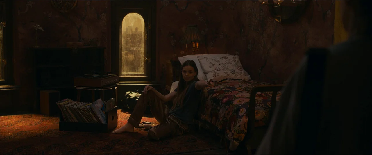 Birds of Paradise (2021) movie still: medium — A dimly lit room contains a bed with floral patterned bedding and records. A woman sits on the floor…