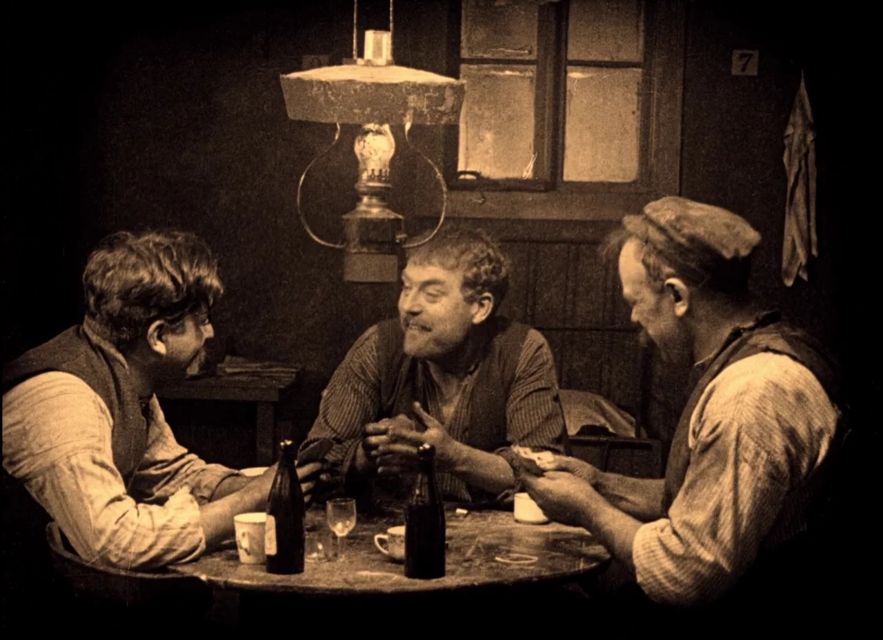 The Phantom Carriage (1921) movie still: medium — A dimly lit room with a single overhead hanging lamp illuminating a round wooden table. Three men…