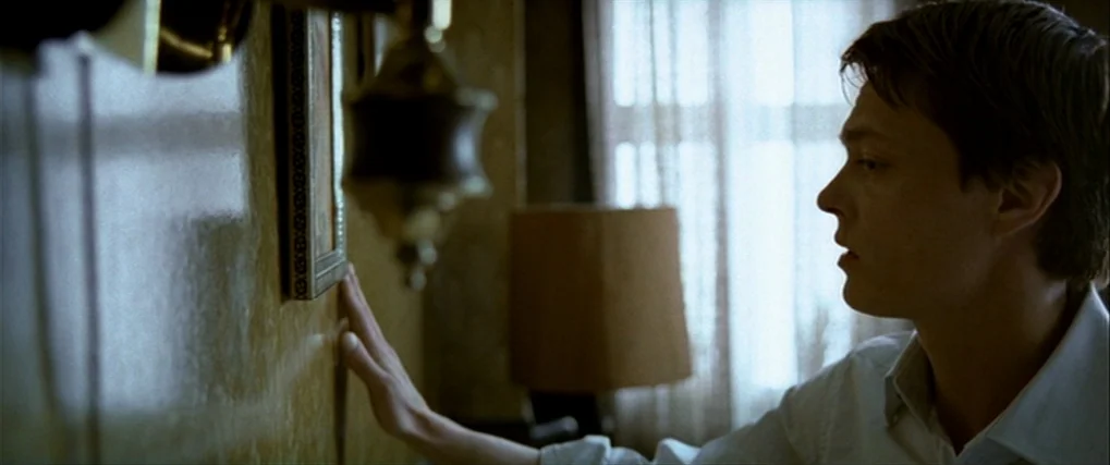 Next Door (2005) movie still: medium — A room with textured yellow wallpaper featuring a wall-mounted framed photograph or object. The subject…