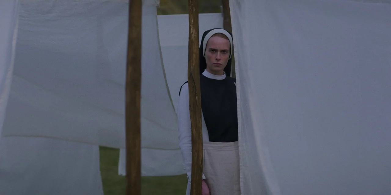 Immaculate (2024) movie still: medium — A woman dressed as a nun stands amidst hanging white laundry sheets on poles. She faces forward while…