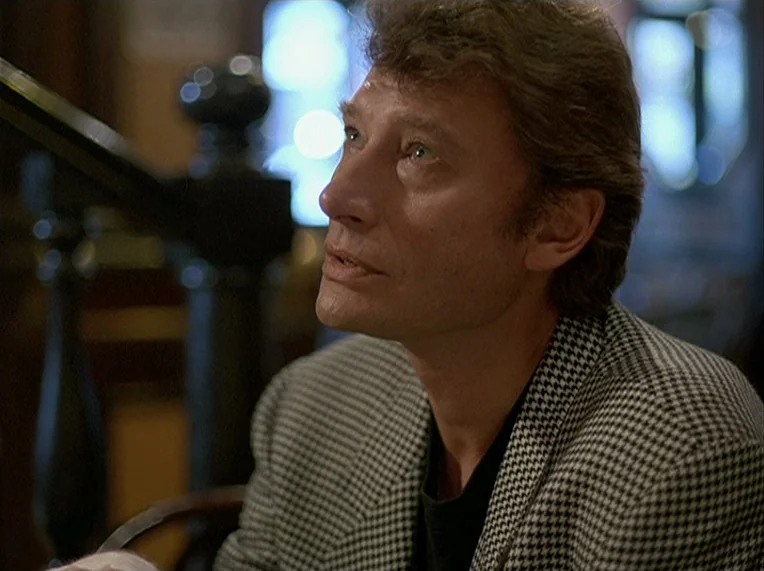 Detective (1985) movie still: closeup — A dimly lit indoor space features blurred architectural details like a wooden staircase railing. A…