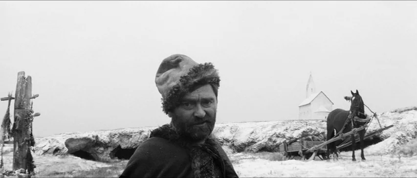 Marketa Lazarová (1967) movie still: medium — A snowy, barren landscape features a distant small white building and a dark horse. The bearded man in…