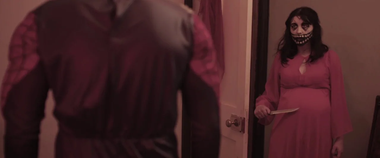 Prevenge (2016) movie still: medium — A woman stands in a dark doorway wearing dark face paint and a dress. She holds a kitchen knife in her right…