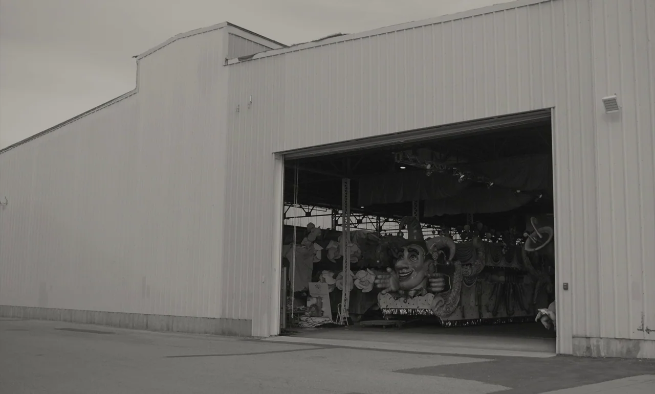 C’mon C’mon (2021) movie still: wide — A large metal industrial shed stands under an overcast sky with an open hangar door. Inside the dark interior…