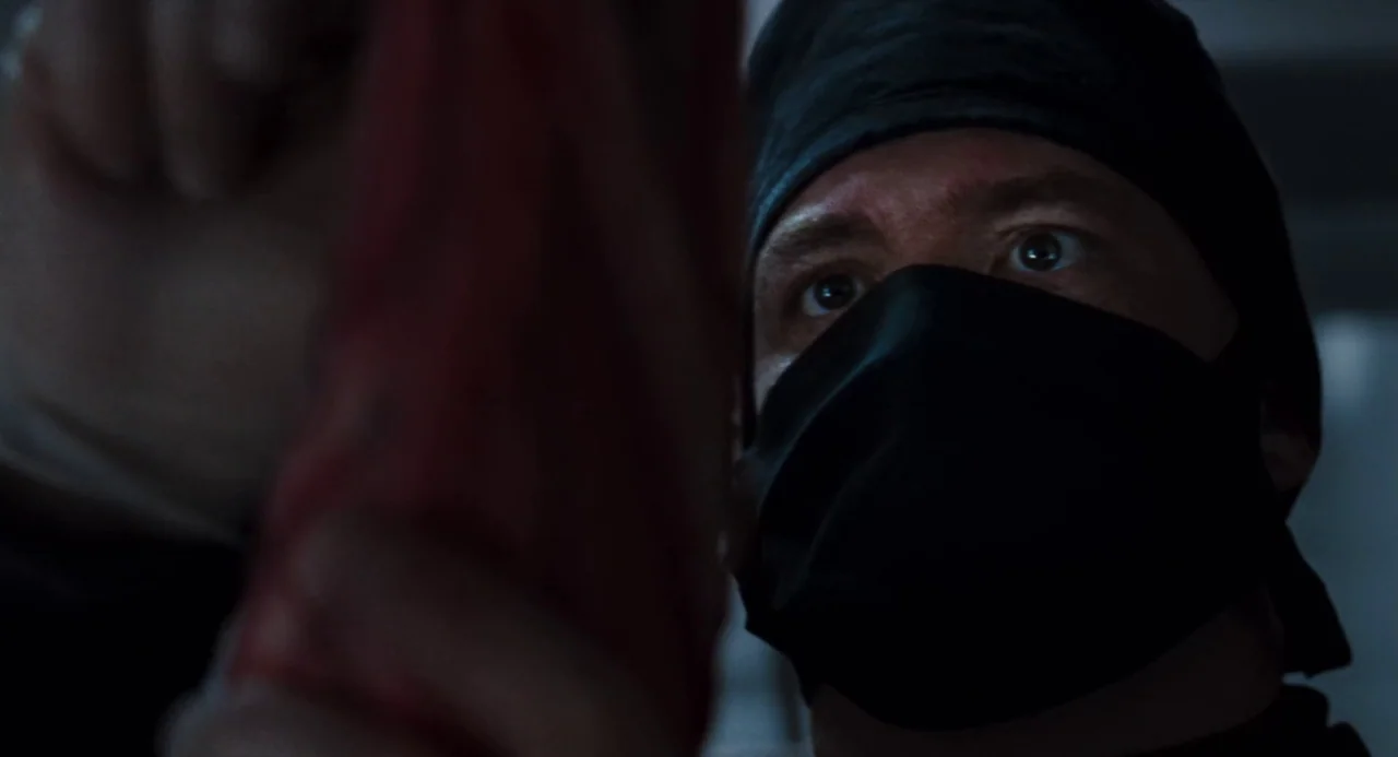 Martyrs (2008) movie still: closeup — A dimly lit room contains a person wearing surgical headgear and a face mask. The individual focuses closely…