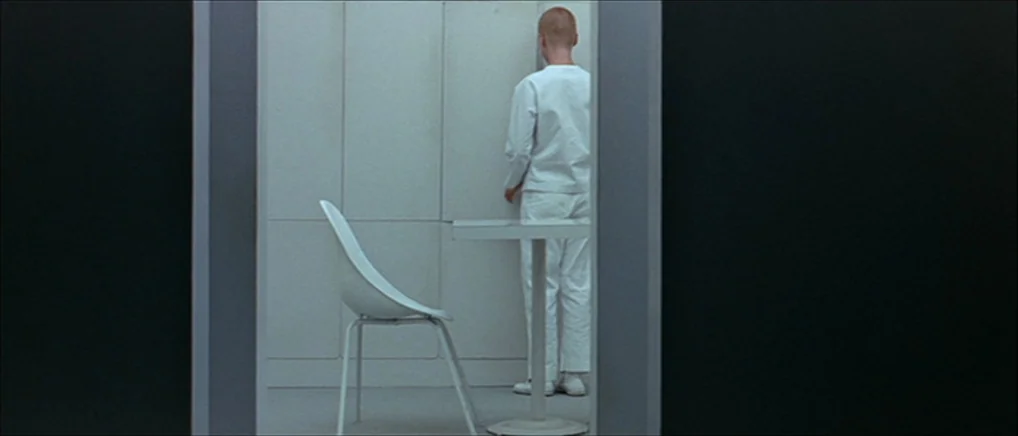 THX 1138 (1971) movie still: wide — A stark white cubicle sits centered between two solid black walls. A person in white clothing stands facing the…