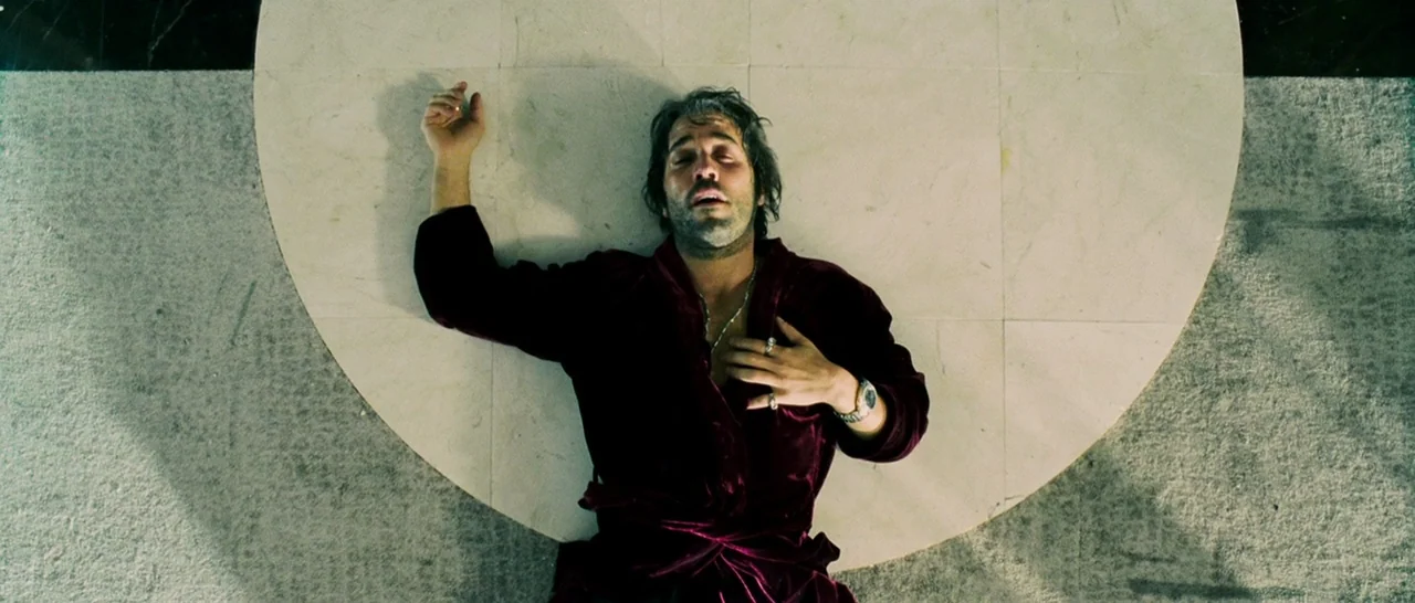 Slice (2018) movie still: fullbody — A man lies supine on a circular marble floor inlay set within a larger floor. The man is wearing a velvet robe…