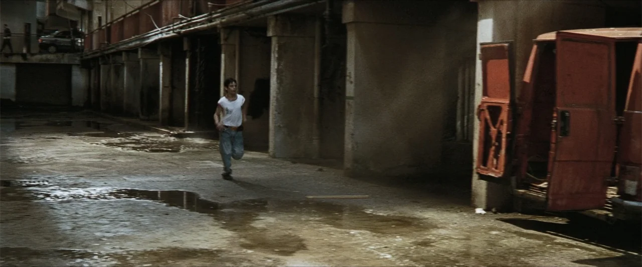 GOMORRAH (2023) movie still: fullbody — A concrete underpass features puddles on the ground and a large orange van parked. A teenage boy in a white…
