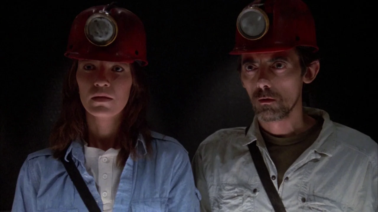 Day of the Dead (1985) movie still: medium — Two individuals stand in a completely dark environment. They both wear red hard hats with active…