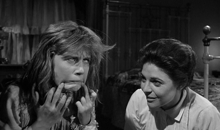 The Miracle Worker (1962) movie still: medium — A dark bedroom interior with a metal bed frame partially visible in the background. A woman observes…