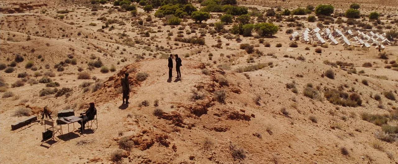 Lord of War (2005) movie still: wide — A dry, arid desert landscape features a rocky ridge overlooking a distant tent encampment. Four men are…