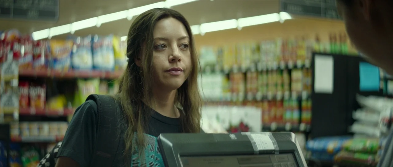 Ingrid Goes West (2017) movie still: medium — A woman stands before a cash register inside a commercial convenience store. Rows of colorful snacks…