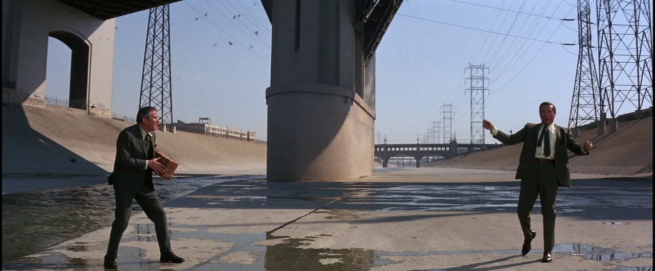 Point Blank (1967) movie still: wide — A wide concrete channel under a bridge with large support pillars and power towers. Two men in identical…
