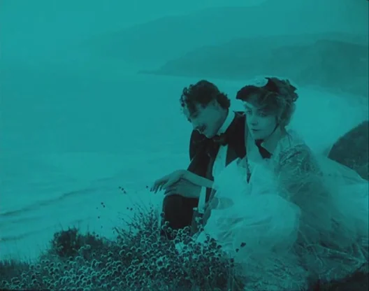 Birth of a Nation (1915) movie still: medium — A man and woman sit together on a high cliff edge overlooking the ocean. The couple is positioned in…