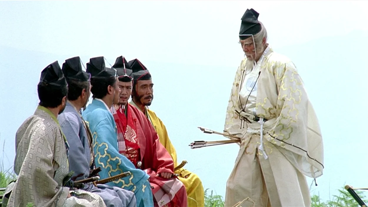 Ran (1985) movie still: fullbody — A group of six men in traditional Japanese attire stand on a grassy ridge. An elderly man in white stands while…