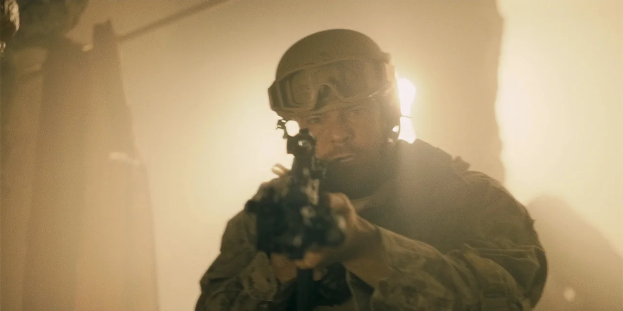 Playdate (2025) movie still: medium — A soldier stands amidst thick dust in a bright, hazy outdoor environment. The man points his rifle forward…