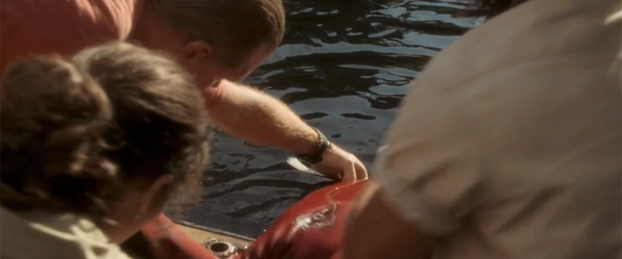 Anaconda (1997) movie still: medium — Three people lean over the side of a boat while examining a submerged object. The dark water reflects the sky…