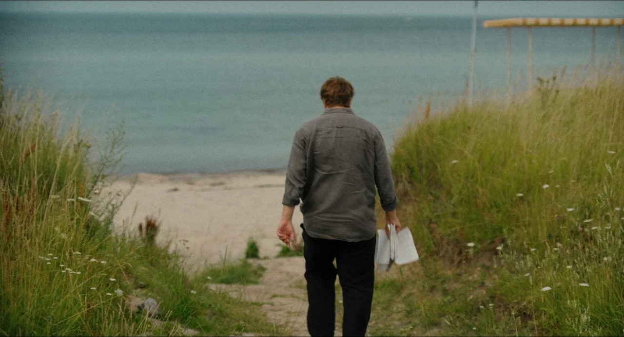 Afire (2023) movie still: fullbody — A man walks down a narrow sand path surrounded by tall green grasses. Beyond the dunes, a stretch of grey water…