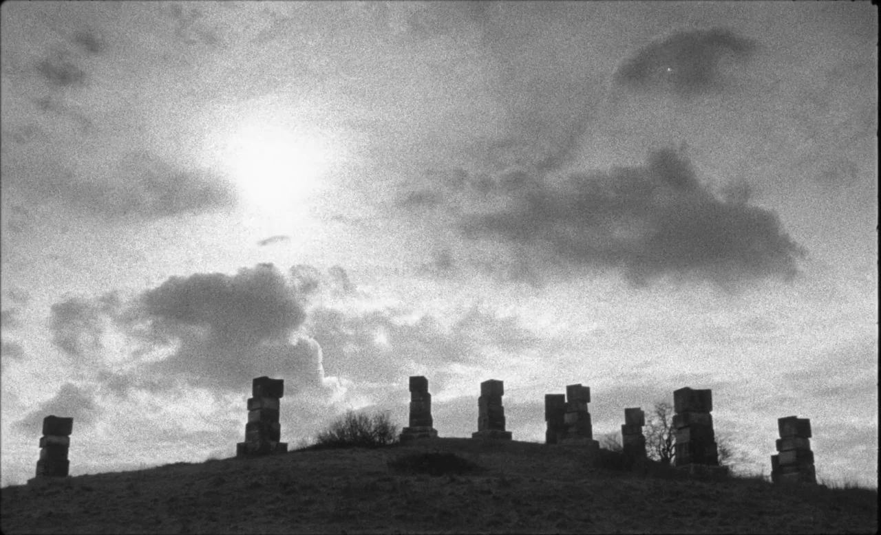 Last and First Men (2020) movie still: wide — A grassy hill features several vertical stone markers against an overcast sky. The sun shines brightly…