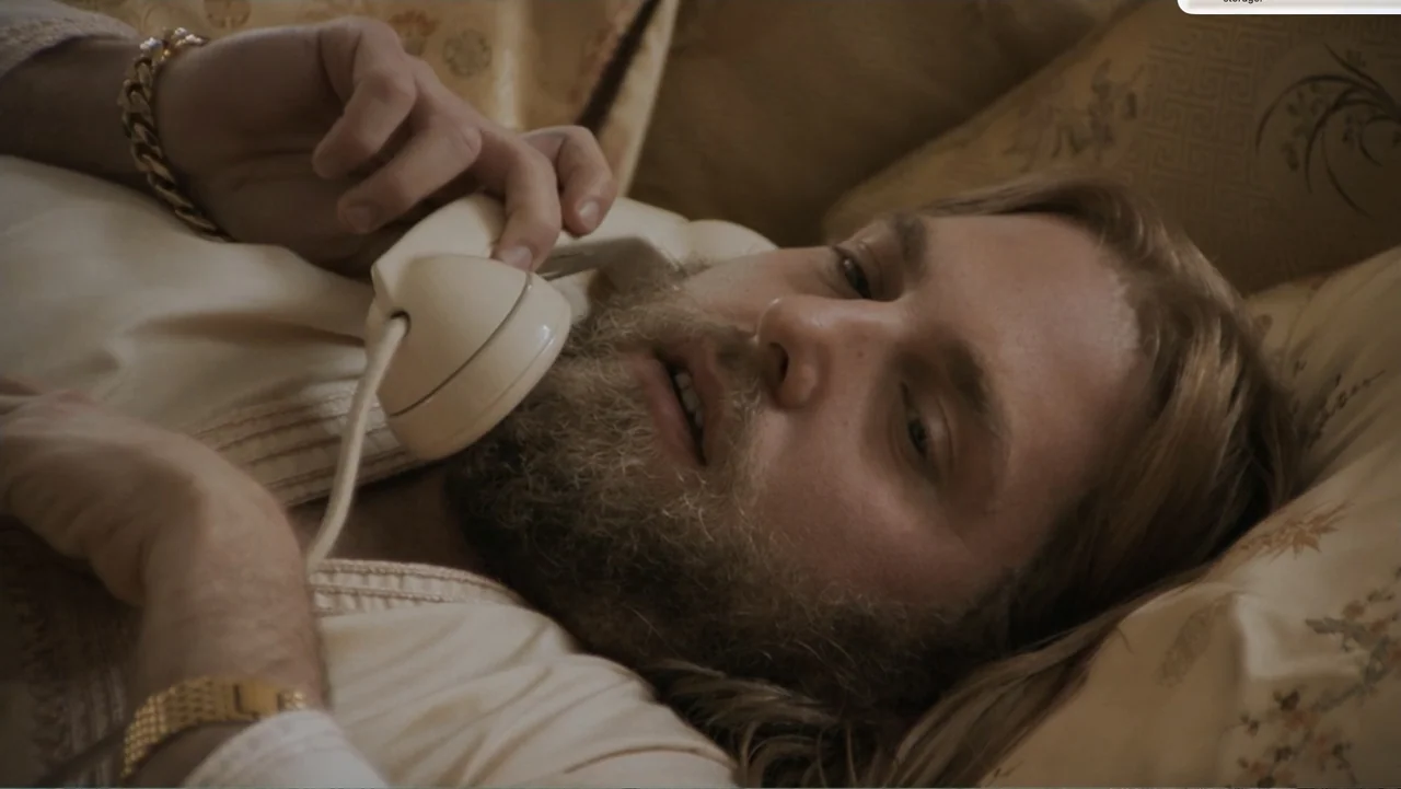 Don't Worry, He Won't Get Far on Foot (2018) movie still: closeup — A man lies on a floral-patterned bed while holding a vintage telephone. He rests…