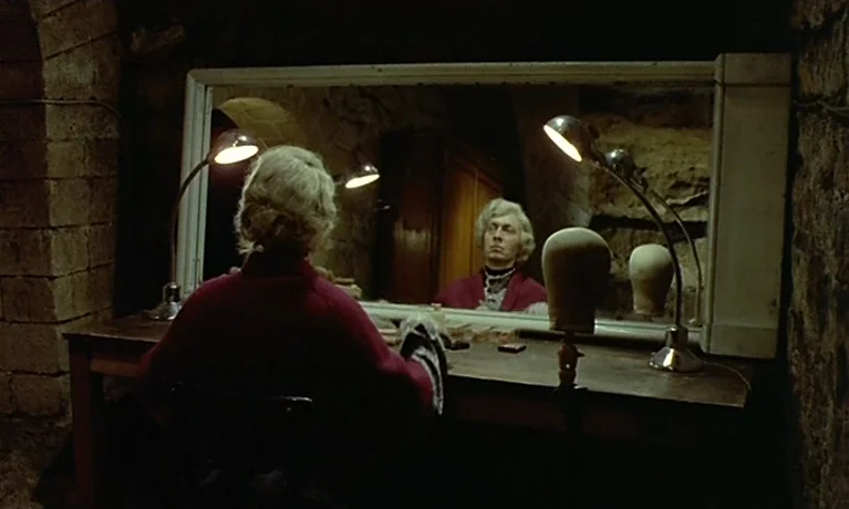 Nuits Rouges (1974) movie still: medium — A stone-walled dressing room features a wooden vanity with a large mirror and lamps. A gray-haired person…