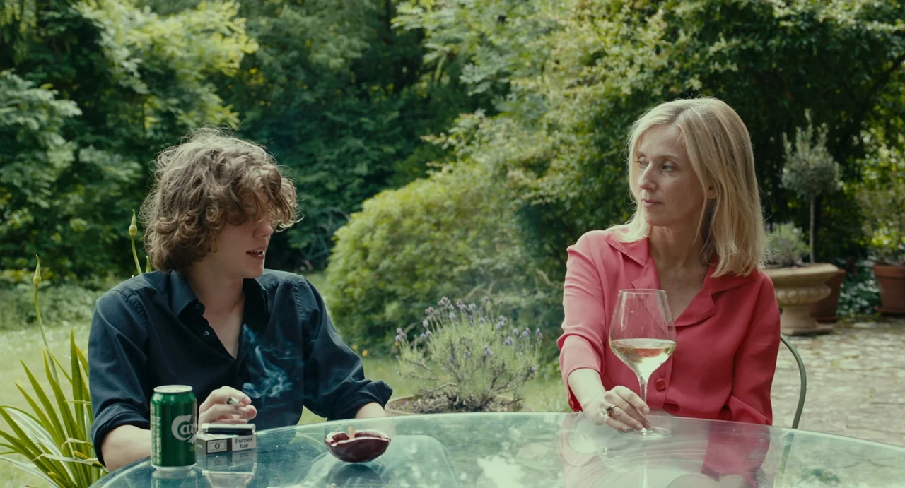 Last Summer (2023) movie still: medium — A glass patio table sits on a paved surface surrounded by lush green foliage. A woman and a young man sit…
