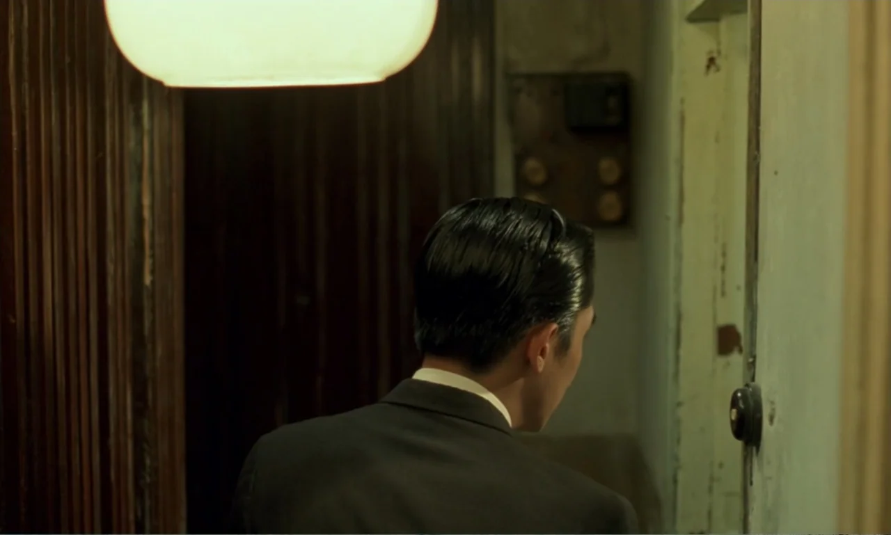In the Mood for Love (2000) movie still: medium — A dimly lit narrow residential hallway features wood-paneled walls and a vintage doorbell. A man…