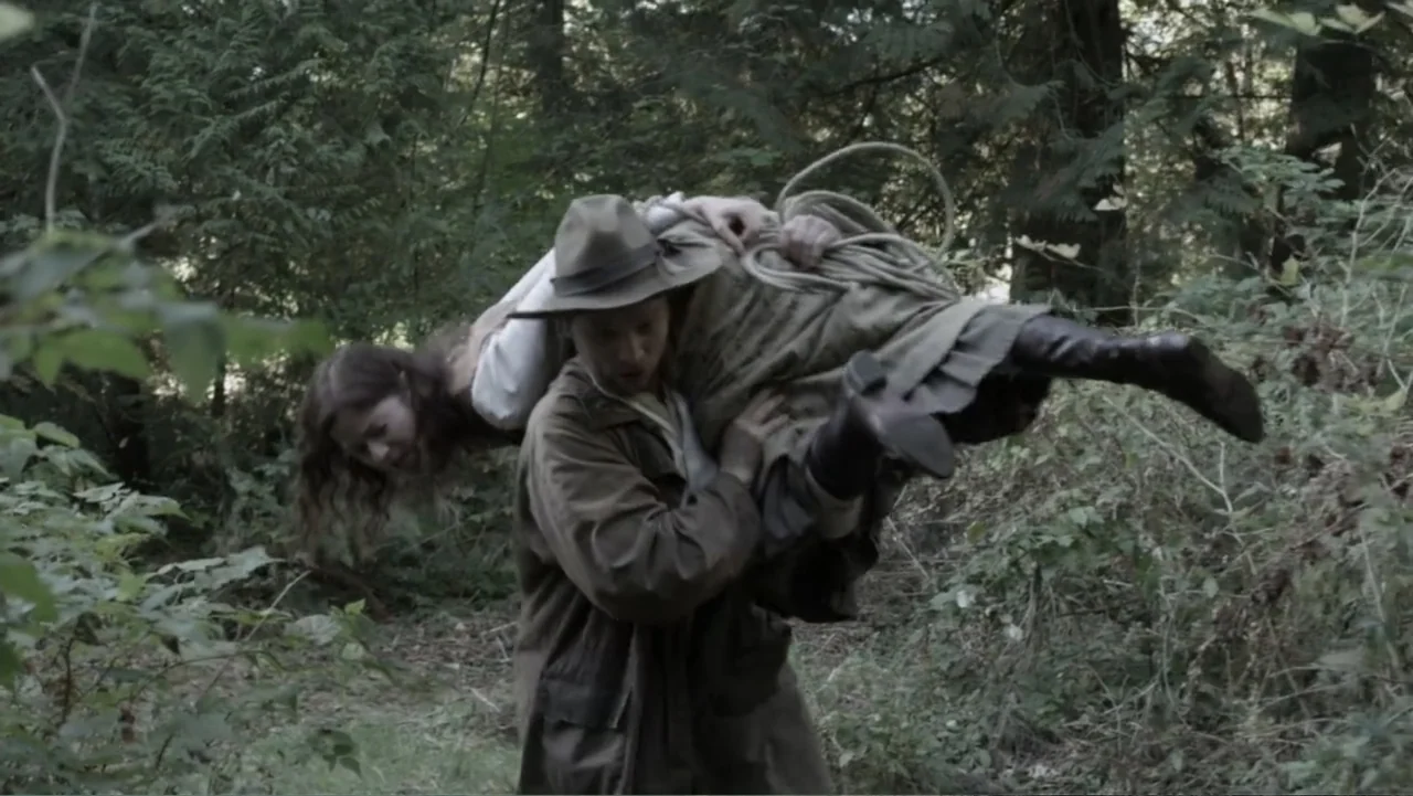 Dawn Rider (2012) movie still: medium — A man in a tan duster walks through a thicket of evergreen trees. He carries a woman horizontally over his…
