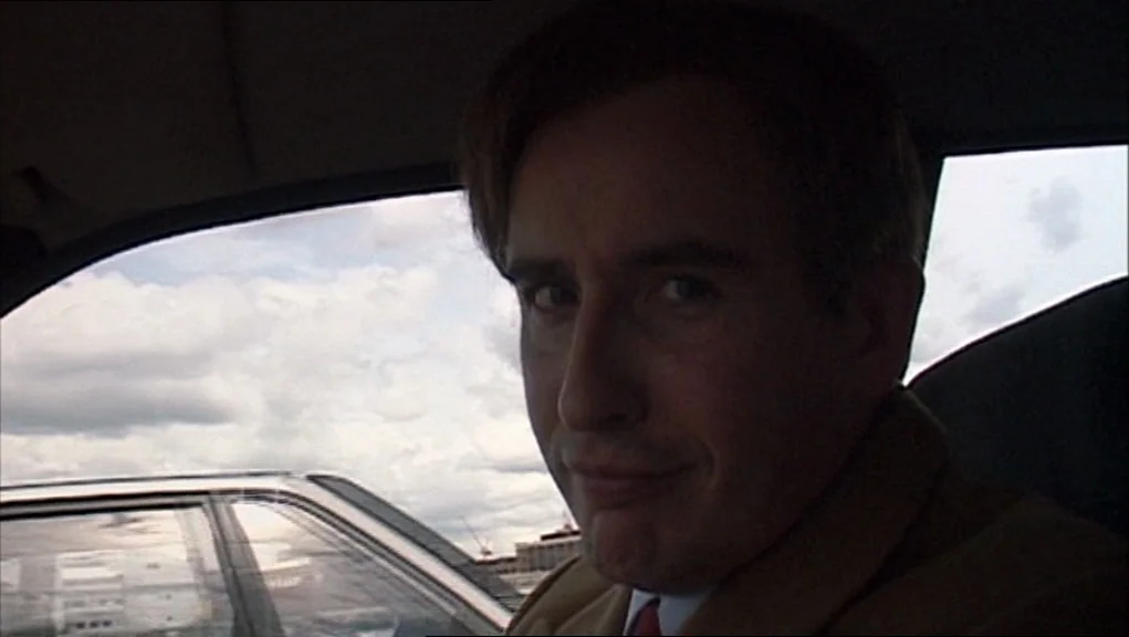 24 Hour Party People movie still: closeup — A man sits inside a vehicle with an overcast sky visible through the windows. The man gazes toward the…