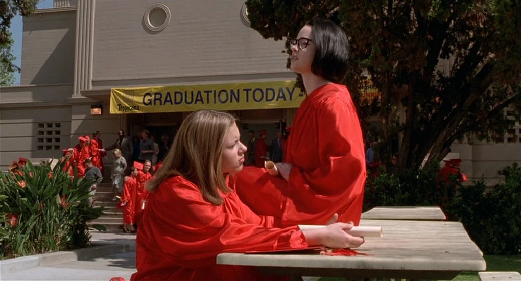 Ghost World (2001) movie still: medium — An outdoor area with a picnic table in front of a school building. Two female students in bright red…