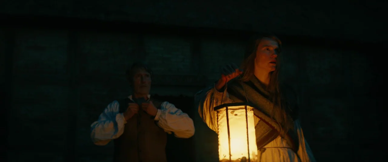 The Promised Land (2023) movie still: medium — Two individuals stand in front of a dark timber-framed structure at night. The woman holds a lantern…