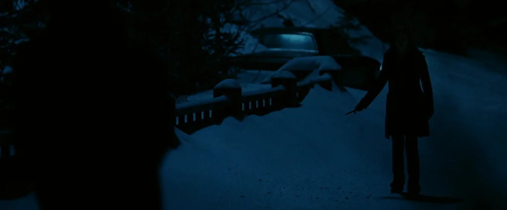 Wind Chill (2007) movie still: wide — A snow-covered road at night is illuminated by dim blue moonlight. A woman holding a small object faces an…