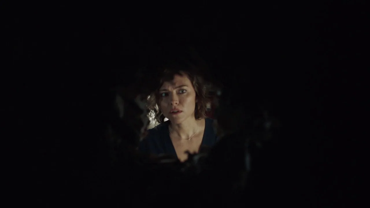 Girl on the Third Floor (2019) movie still: closeup — A woman stares through an irregular hole in a dark, shadow-filled partition. Her face is the…