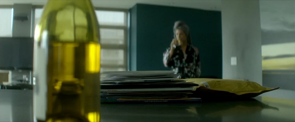 Haywire (2011) movie still: wide — A modern living space features a countertop foreground with a bottle and mail pile. A woman in a robe stands…
