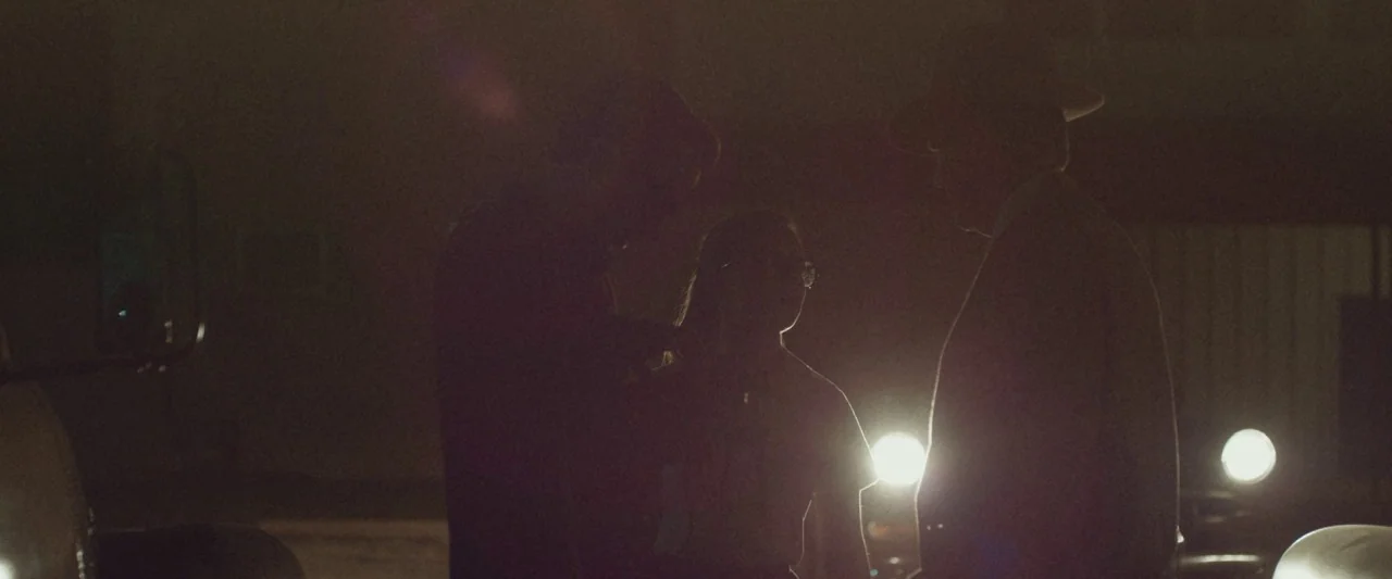 The Vast of Night (2019) movie still: medium — An outdoor location at night with three figures standing in dark silhouettes. Bright vehicle…