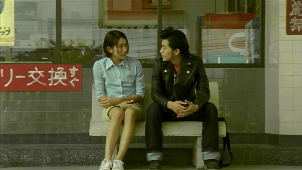 Wild Zero (1999) movie still: medium — A tiled storefront exterior features a concrete bench against a large glass window. A young woman and a man…