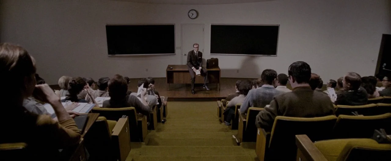 A Single Man (2009) movie still: wide — A mid-century modern lecture hall with curved white walls and yellow tiered seating. A man in a suit sits on…