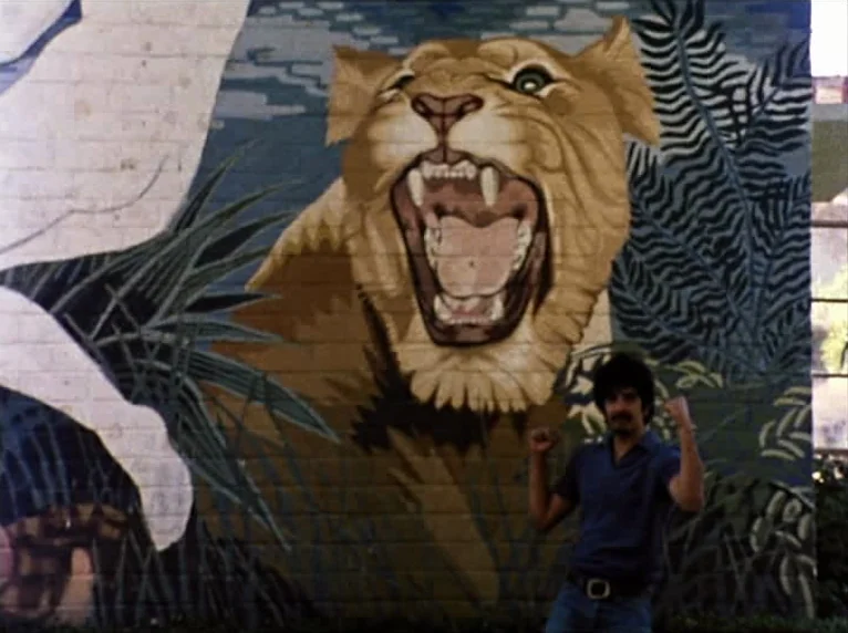 Mur Murs (1981) movie still: medium — A brick wall features a large, colorful painted mural of a roaring lion. A man stands in front of the wall and…