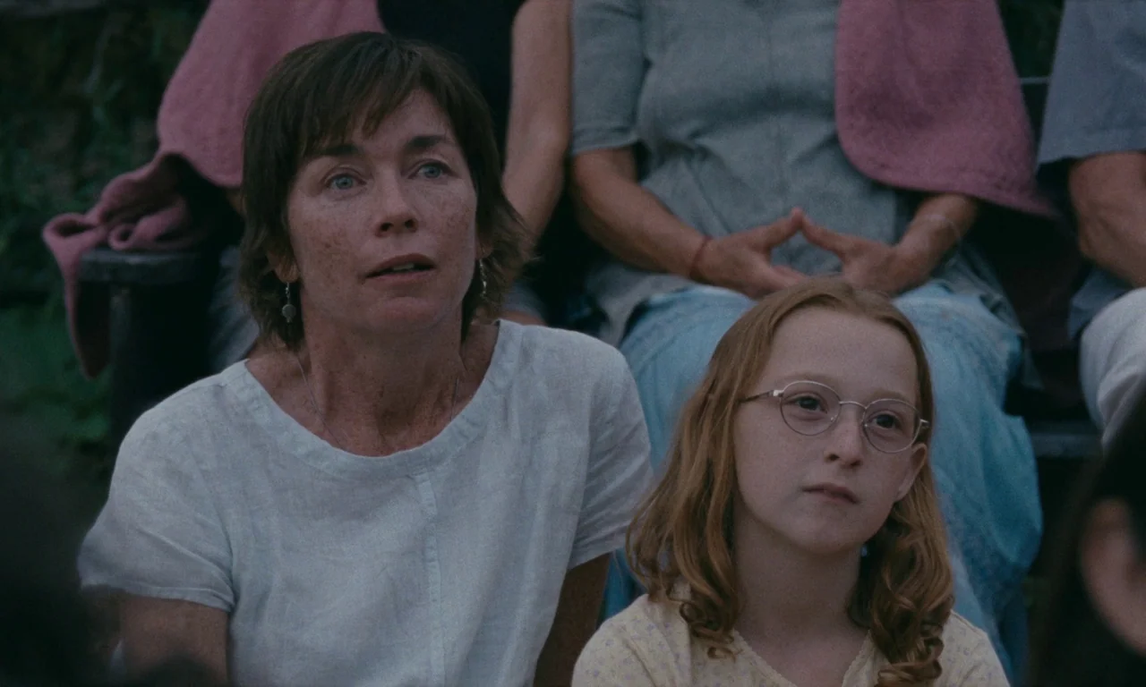 Janet Planet (2023) movie still: medium — An outdoor seating area with people partially visible behind a woman and a child. The pair sits together…