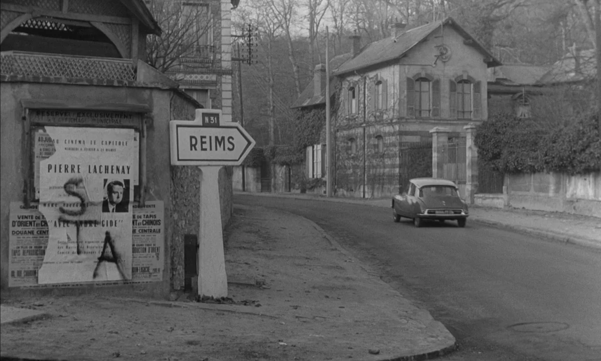 La Peau Douce (The Soft Skin) (1964) movie still: wide — A quiet road winds past a stone wall with torn posters and a road sign. A single vintage…